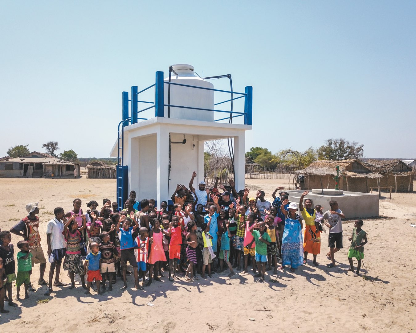 A large group of Malagasy men, women, and children smiling and waving in front of a water tower in their village