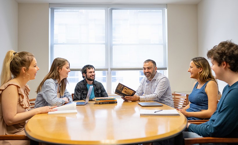 Students sit around a table with a professor in discussion.